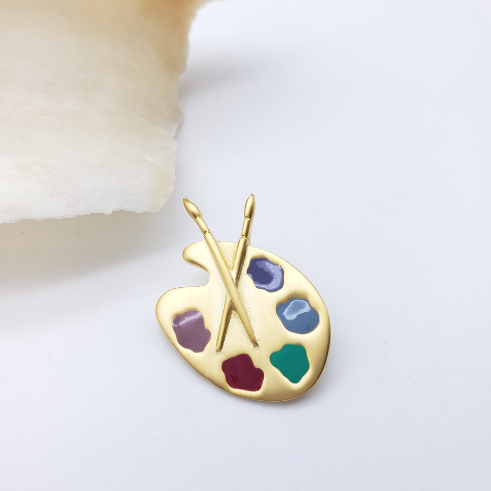 A gold-colored enamel pin shaped like an artist's paint palette with paintbrushes, featuring multicolor enamel paint spots on a white background.