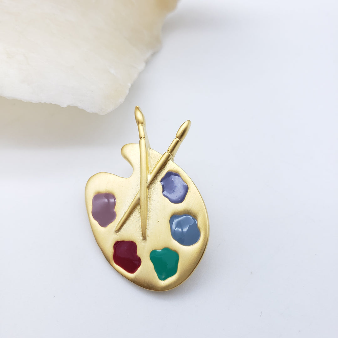 A gold artist's paint palette brooch with six painted color wells and two crossed paintbrushes, photographed on a clean white surface with a soft cream stone prop at the corner.