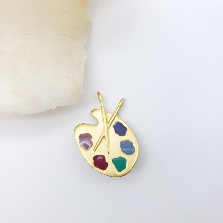 A gold-plated artist's paint palette brooch with colorful enamel paint spots and crossed paintbrushes, displayed on a clean white surface next to a textured cream stone.