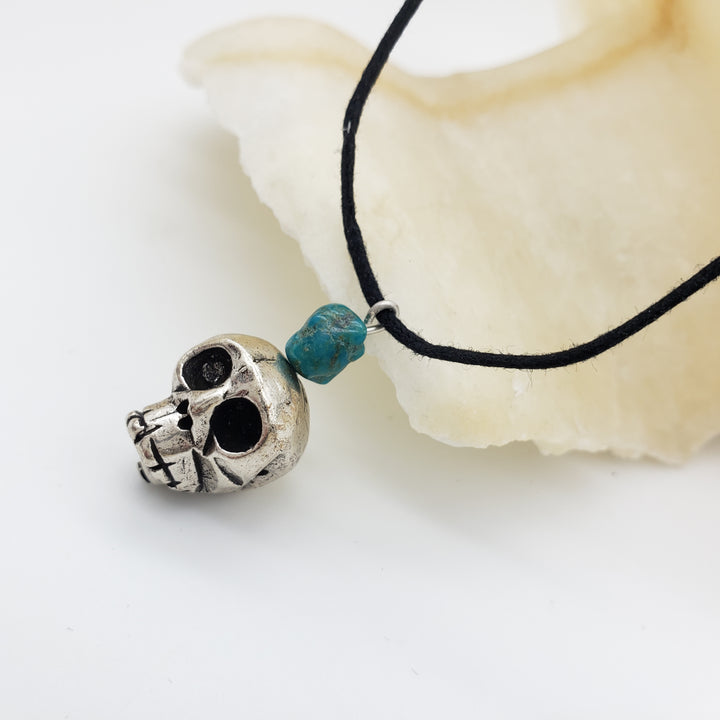 Silver skull pendant with a turquoise bead on a black cord, lying on a white shell.