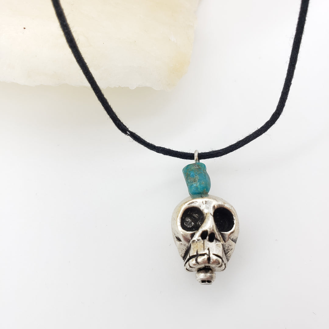 Skull-shaped pendant with a turquoise stone on a black cord against a white background