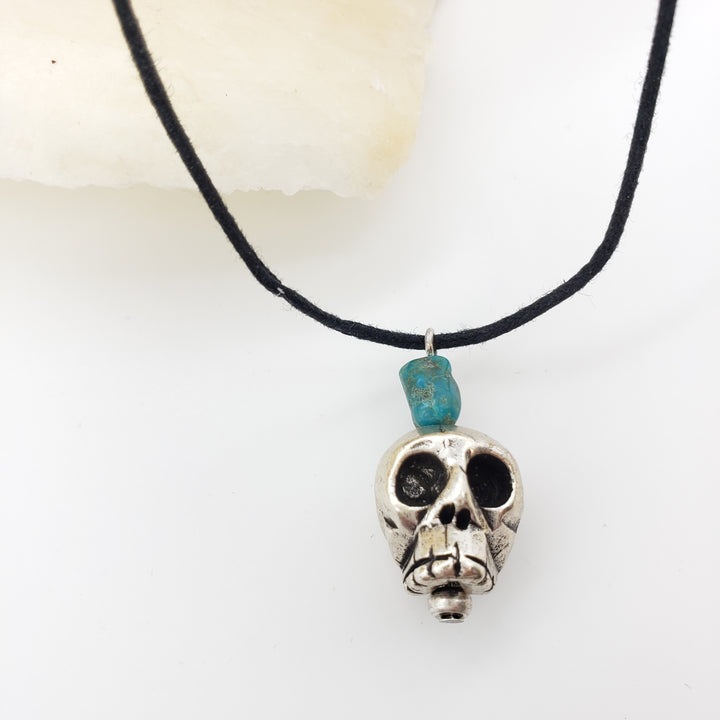 Skull-shaped pendant with a turquoise stone on a black cord against a white background