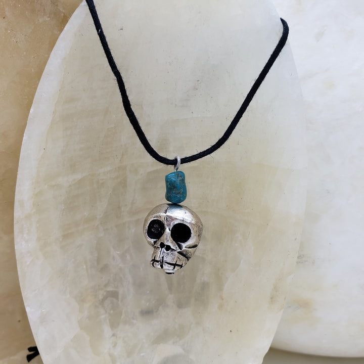 Skull-shaped pendant with turquoise bead on black cord against a stone background