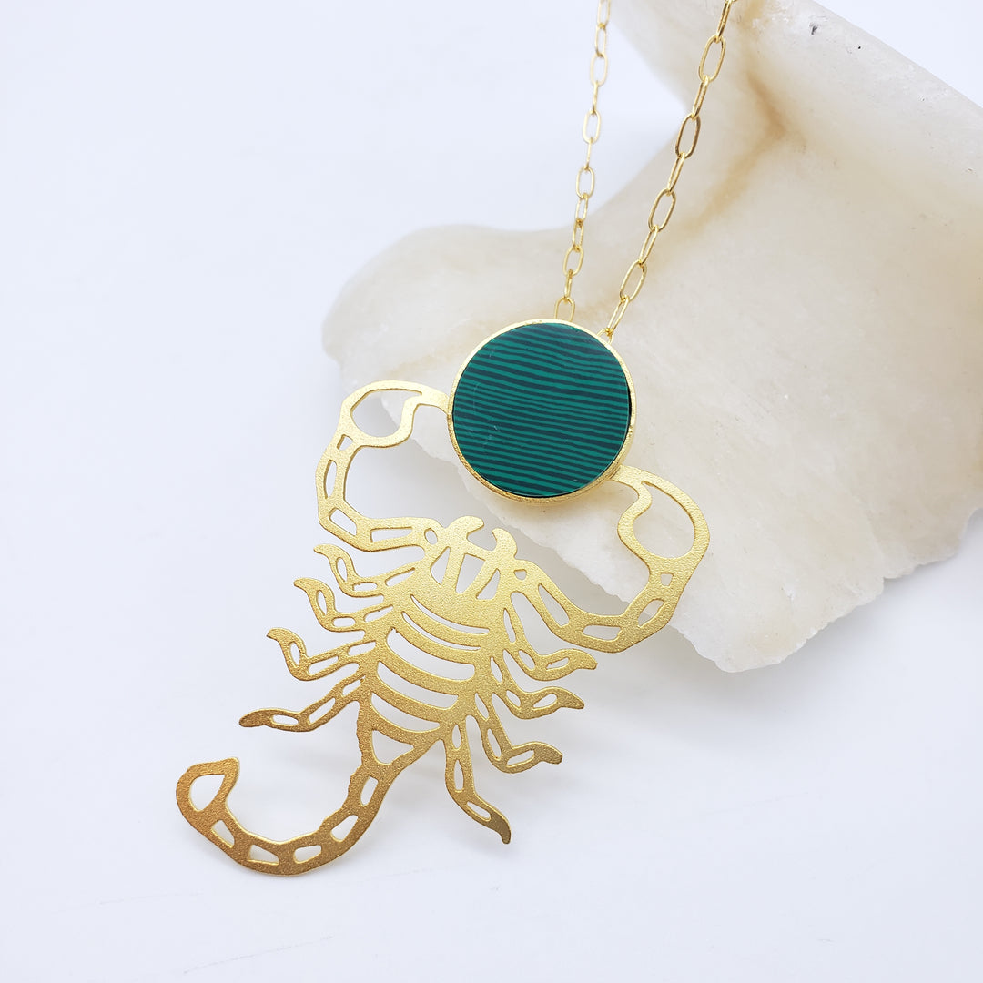 Gold scorpion-shaped pendant with a green circle on a white background