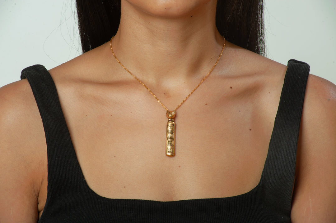Gold necklace with a cylindrical pendant worn by a person against a neutral background