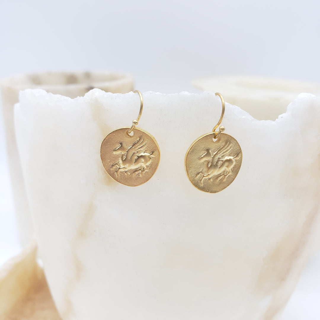 Two gold circular earrings with engraved designs on a white marble surface.