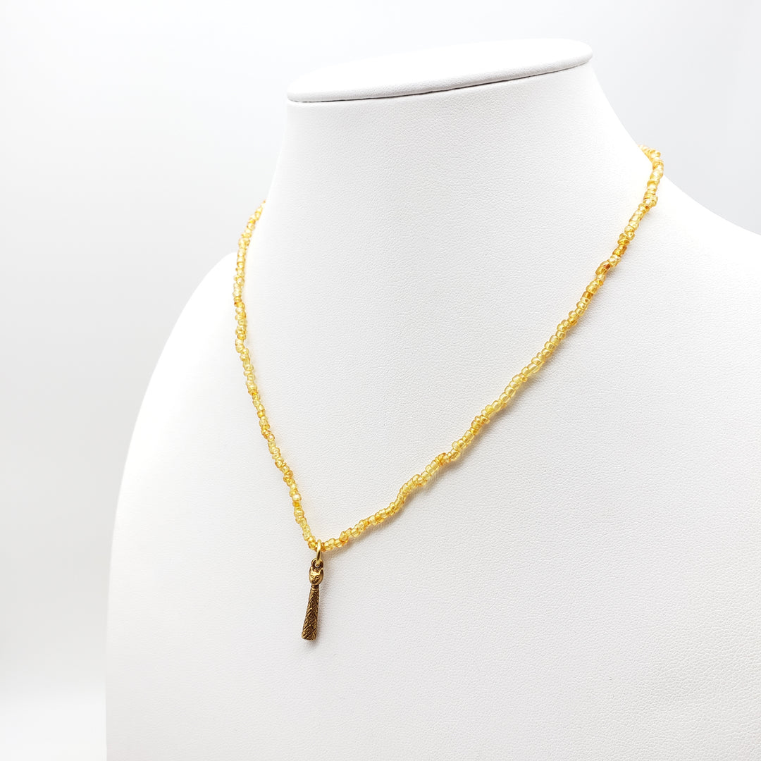 A gold beaded necklace with a small textured gold pendant, displayed on a white mannequin bust against a white background.