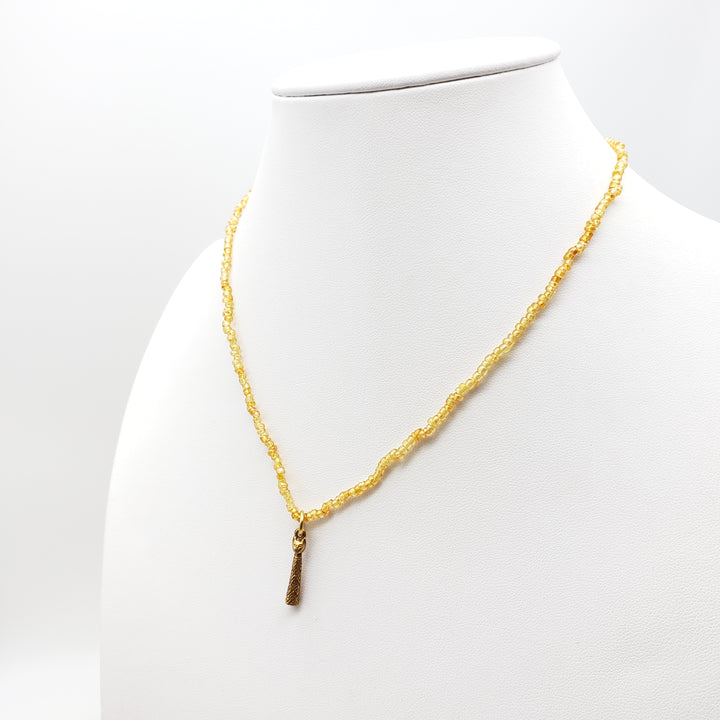 A gold beaded necklace with a small textured gold pendant, displayed on a white mannequin bust against a white background.