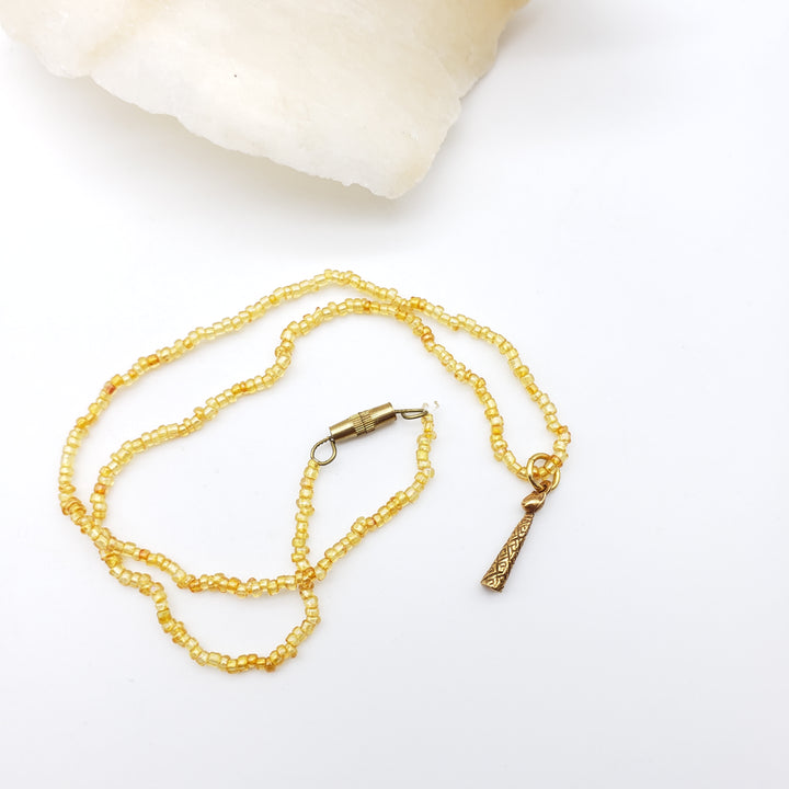 A delicate gold beaded necklace with a small engraved pendant, displayed on a clean white surface with a white crystal shell in the background.