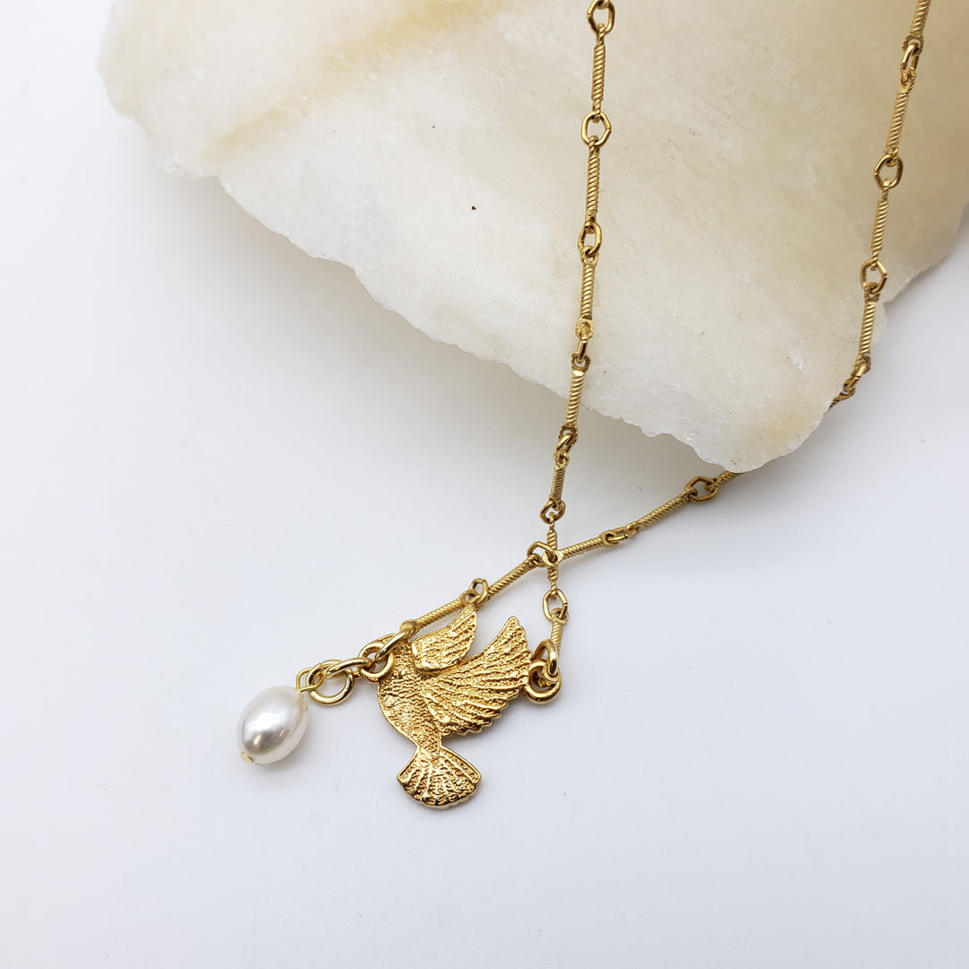 A gold-tone chain necklace with a bird-shaped pendant and a dangling pearl, displayed on a white surface next to a polished stone.