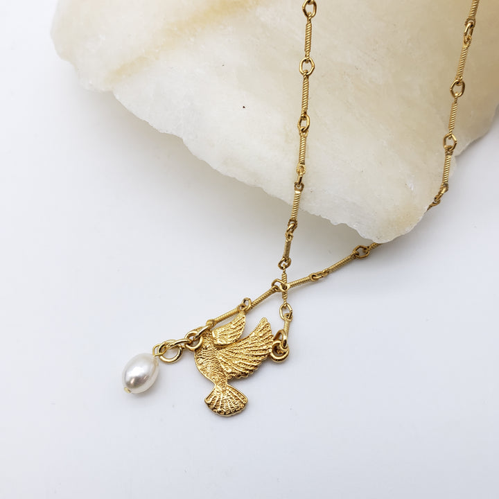 A gold-tone chain necklace with a bird-shaped pendant and a dangling pearl, displayed on a white surface next to a polished stone.