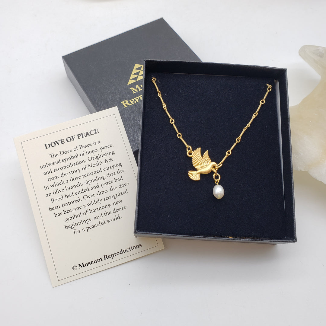 A gold-tone dove necklace with a white pearl pendant displayed in an open black presentation box, alongside a branded black box lid and a printed card titled 'DOVE OF PEACE' on a white background.