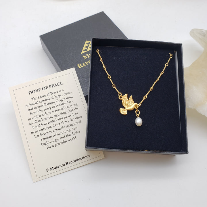 A gold-tone dove necklace with a white pearl pendant displayed in an open black presentation box, alongside a branded black box lid and a printed card titled 'DOVE OF PEACE' on a white background.