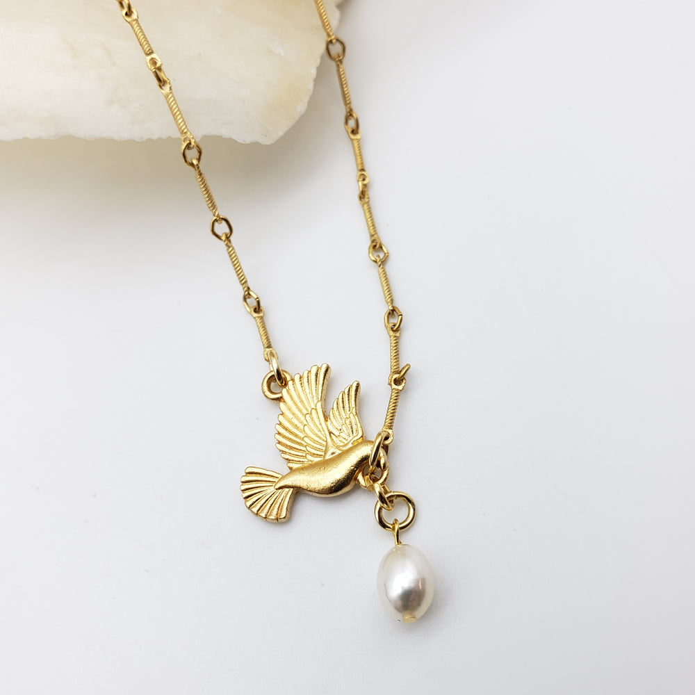 Gold-tone necklace featuring a bird pendant with a dangling white pearl, displayed on a clean white surface with a small decorative stone.