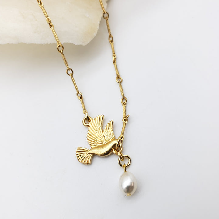 Gold-tone necklace featuring a bird pendant with a dangling white pearl, displayed on a clean white surface with a small decorative stone.