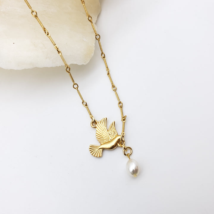 A gold-toned chain necklace featuring a small bird pendant and a dangling white pearl, displayed on a clean white surface with a light stone prop.