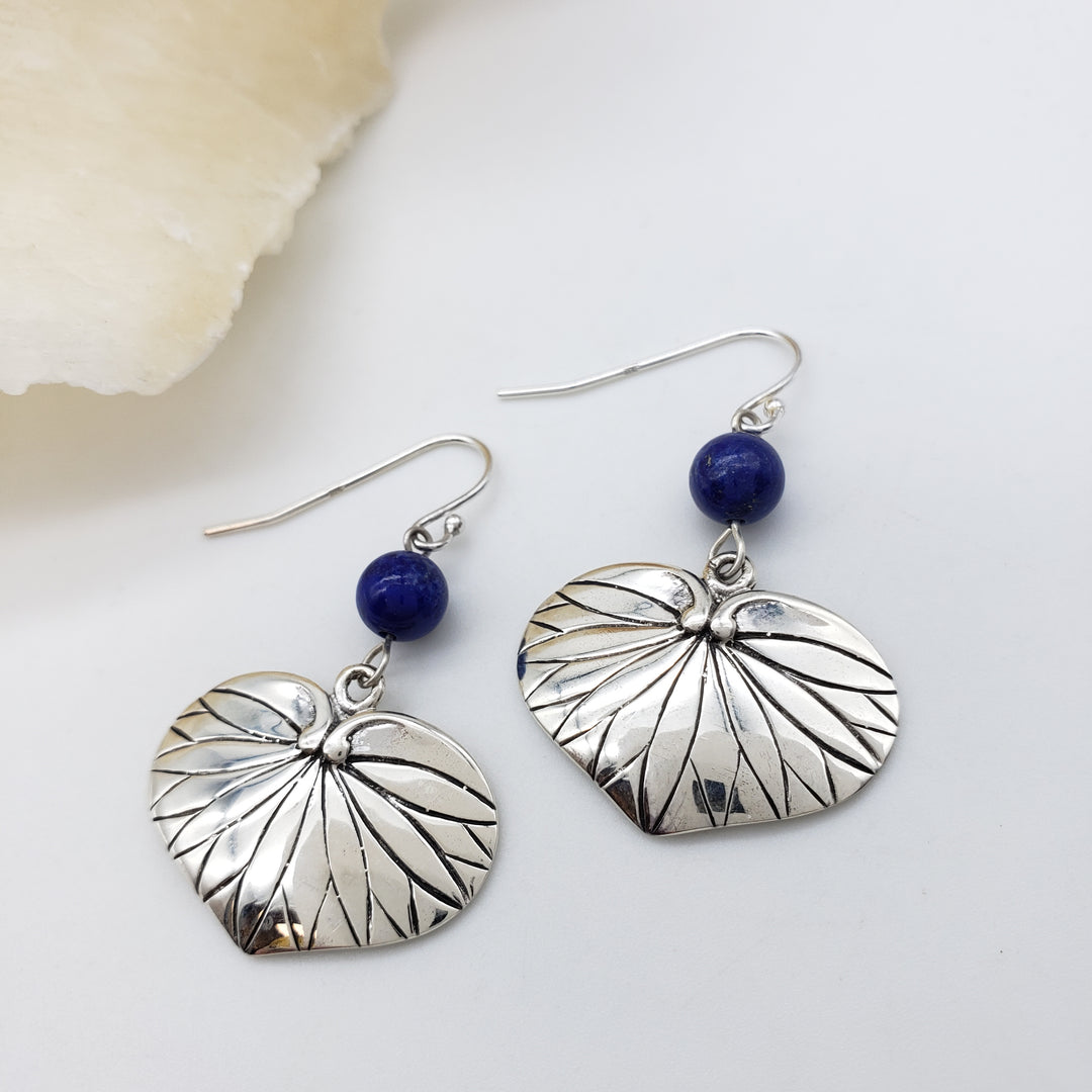 A pair of silver dangle earrings with textured, leaf-like heart-shaped charms and a small round dark blue bead near the hooks, displayed on a clean white surface.