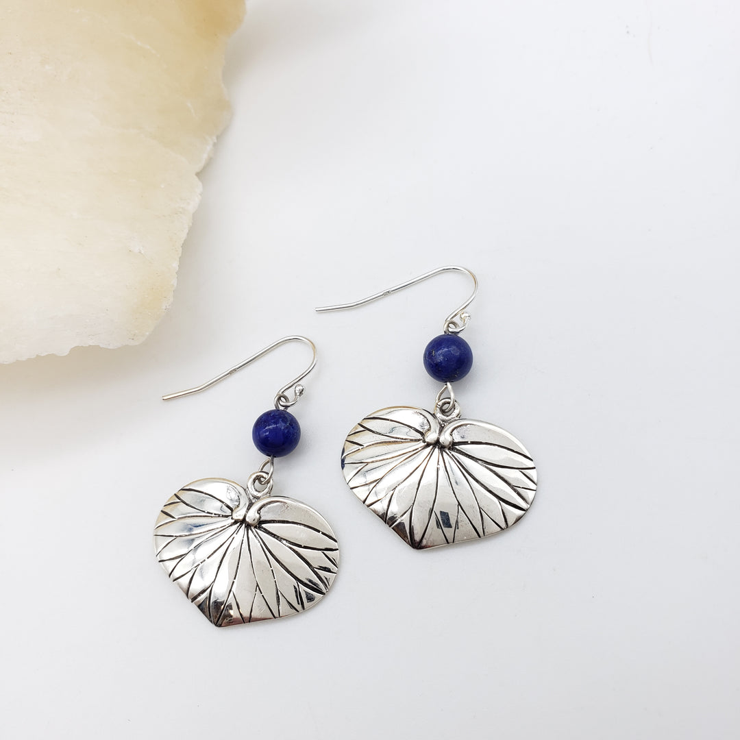 A pair of silver dangle earrings with textured leaf-shaped charms and small dark blue beads, displayed on a white surface next to a pale stone slice.