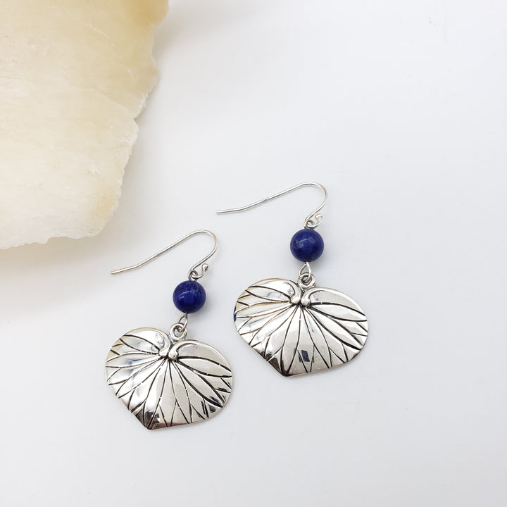 A pair of silver dangle earrings with textured leaf-shaped charms and small dark blue beads, displayed on a white surface next to a pale stone slice.