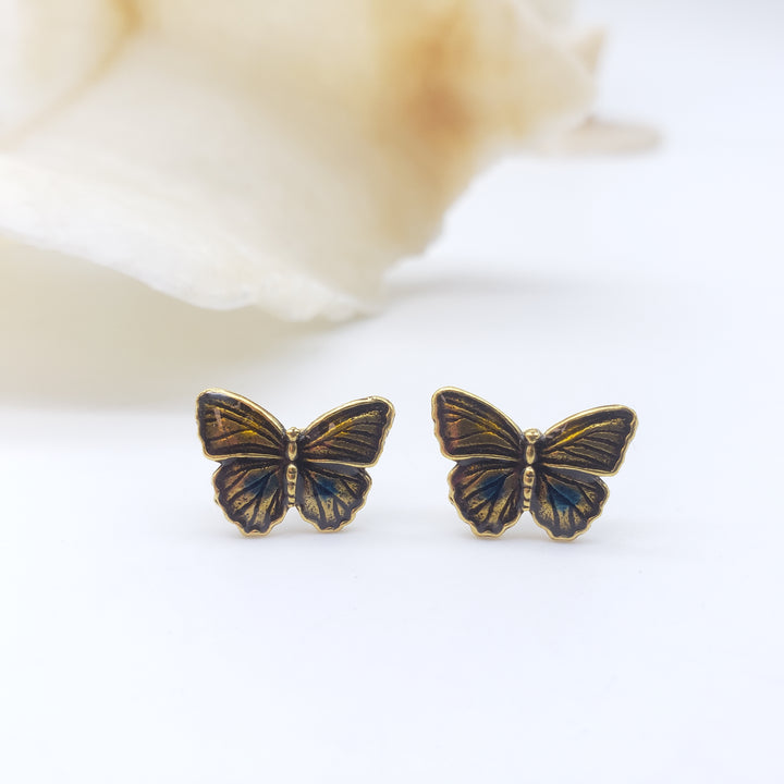 A pair of small butterfly-shaped stud earrings with gold-tone metal and dark blue iridescent enamel wings, displayed on a white surface with a soft blurred cream flower in the background.