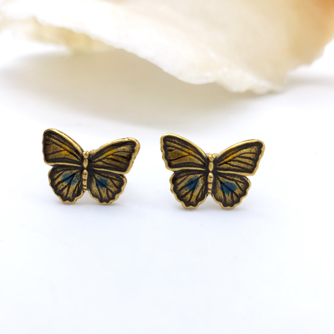 A pair of butterfly stud earrings with gold-toned metal and blue-green enamel detailing, displayed on a clean white surface with a soft-focus seashell in the background.