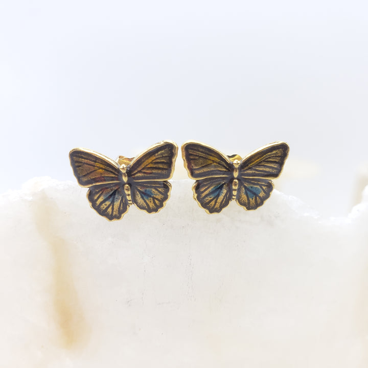 A pair of butterfly-shaped stud earrings with gold-toned metal borders and iridescent blue, green, and bronze wing inlay, resting on a white textured surface against a soft white background.
