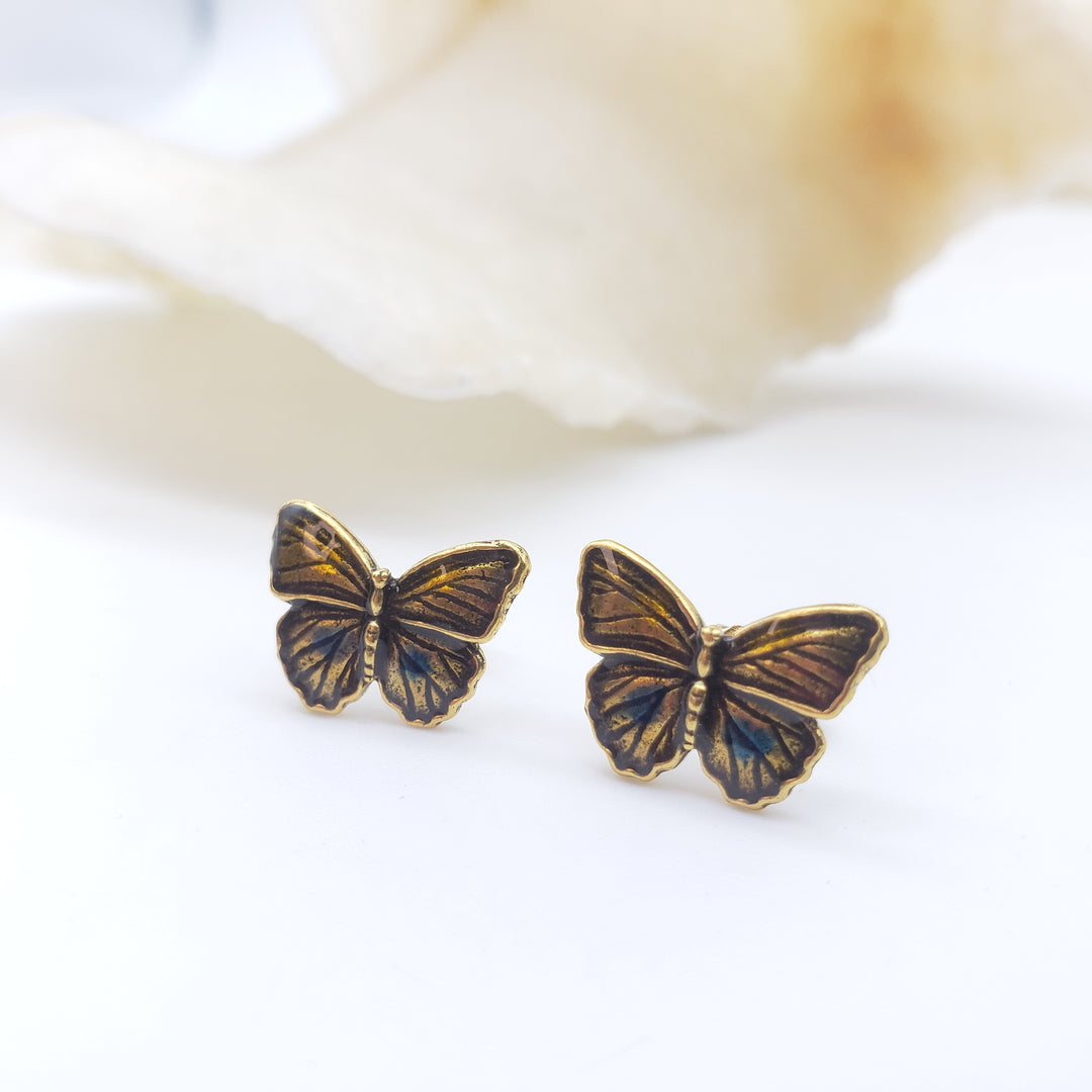A pair of antique bronze butterfly stud earrings with iridescent blue-green wing details, displayed on a clean white surface with a soft-focus beige flower in the background.
